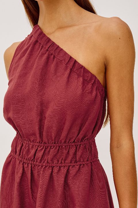 Woman in a spice-colored linen one-shoulder dress with elastic waist and textured fabric, suitable for casual wear.