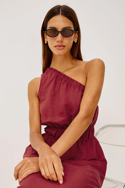 Woman in a spice-colored linen dress sits on a chair, sunglasses on, hands resting in her lap.