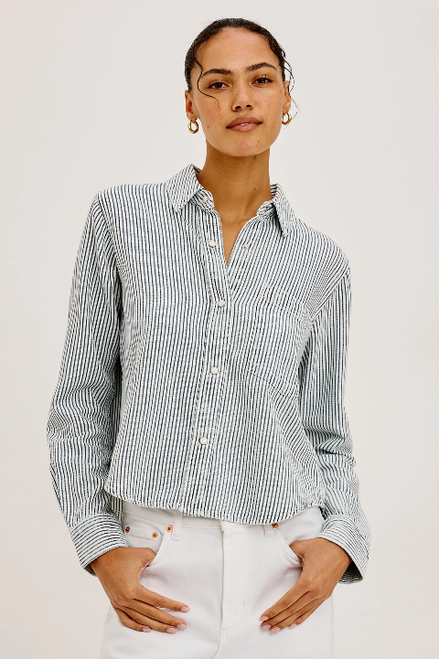Woman in striped button-up shirt and white pants, hands in pockets. Shirt features blue and white railroad stripes.