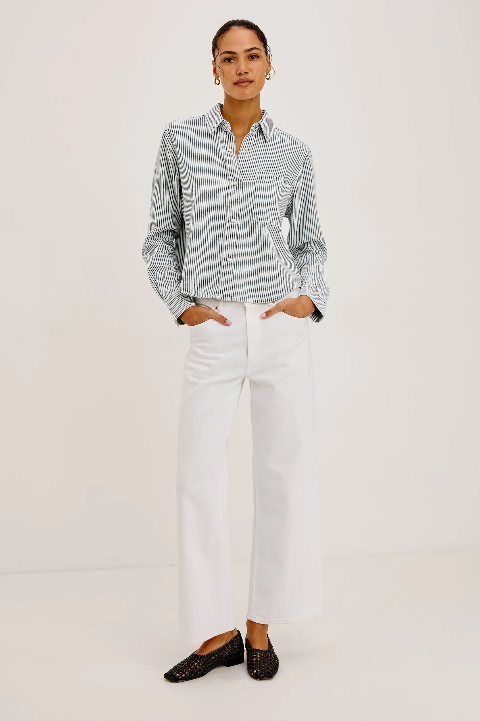 Woman in striped button-up shirt, white wide-leg pants, and black loafers with hands in pockets.
