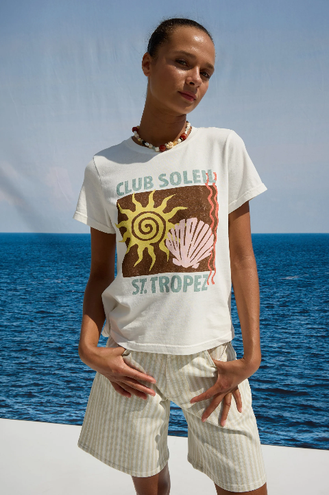 Woman wears a light crewneck sweater, hands in pockets, standing by the ocean with a clear blue sky overhead.