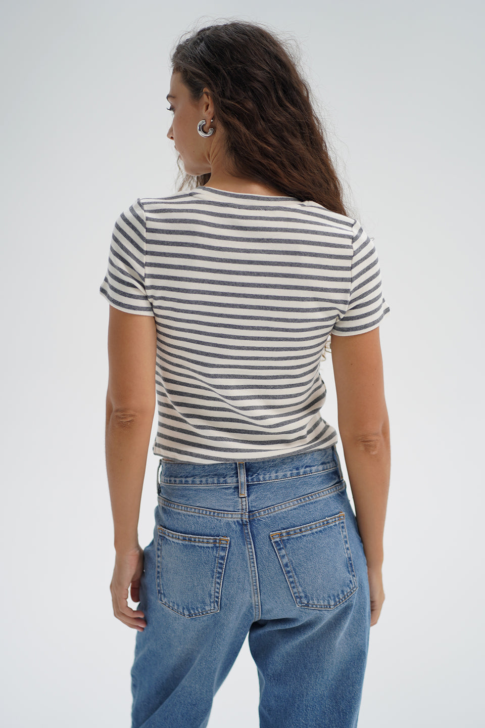 Person with long wavy hair, seen from the back in a greyscale striped cotton tee and blue denim jeans.