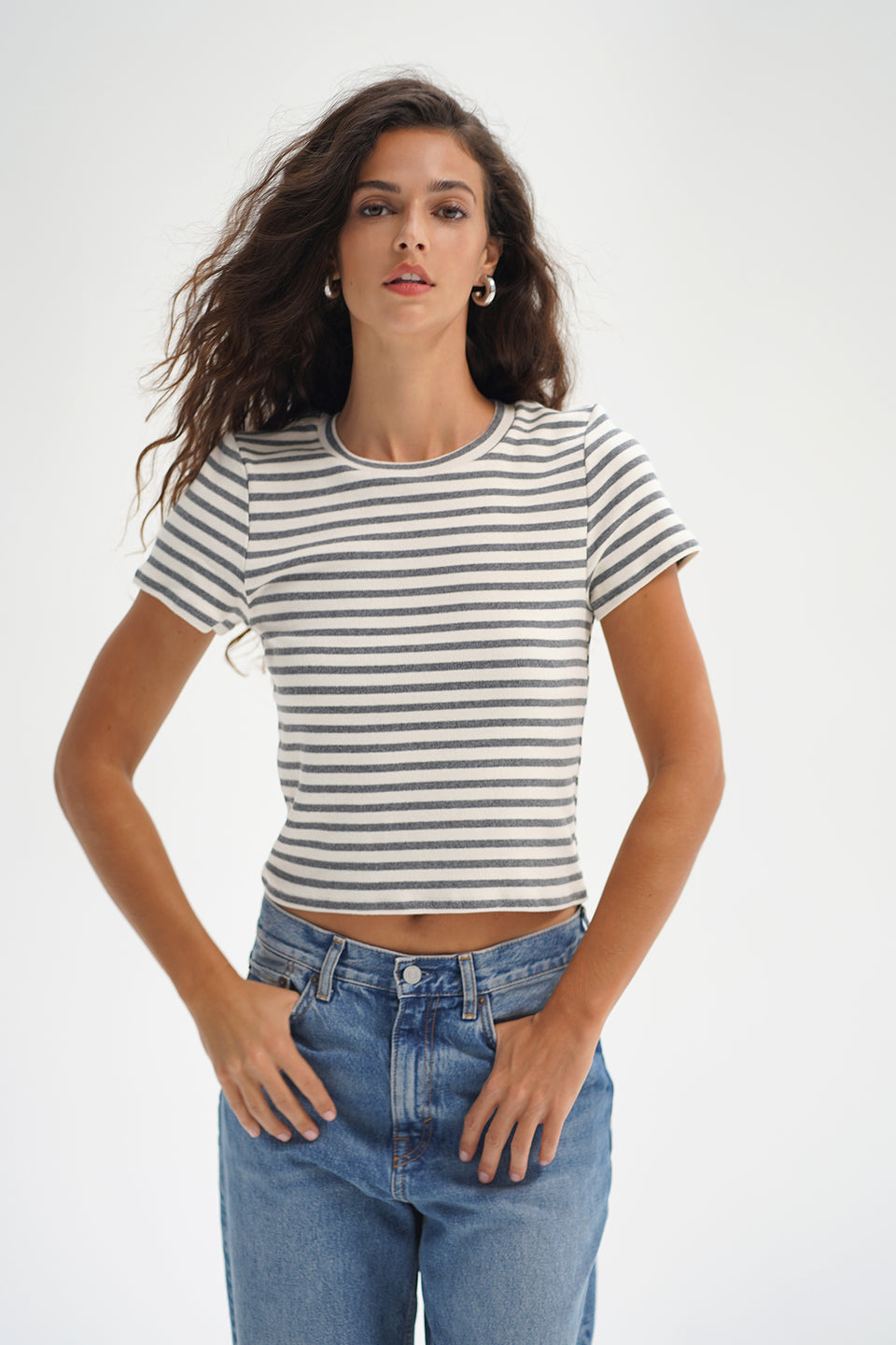 Woman wearing a greyscale striped cotton tee and jeans, standing against a white background, facing forward.