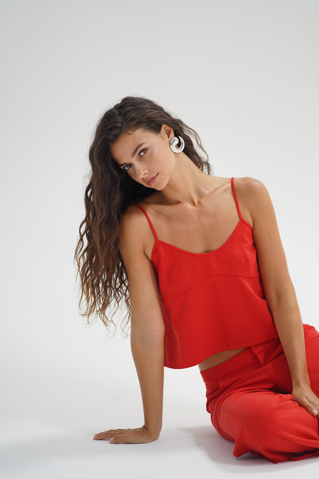 Woman with long wavy hair in a red chambray tank top, seated on the floor against a white background.