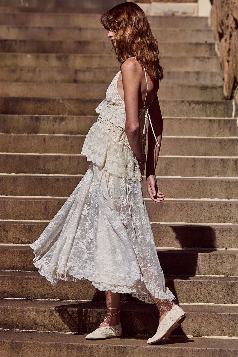 Woman wearing a cream lace maxi skirt and lace-up shoes descends sunlit stone steps.