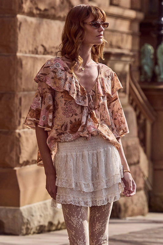 Woman in sunglasses wears a floral ruffled top and cream lace mini skirt, standing outdoors.