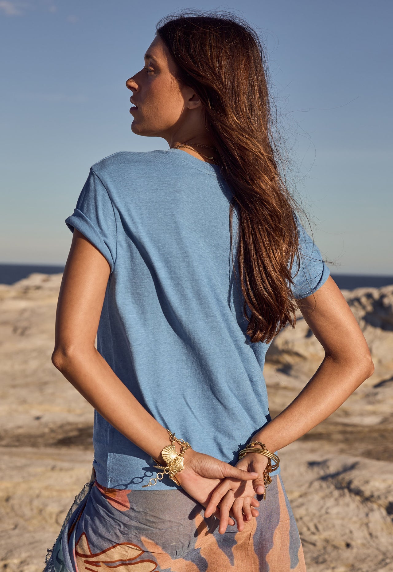 Woman with long brown hair wears a blue cotton tee outdoors, hands clasped, facing a rocky landscape.
