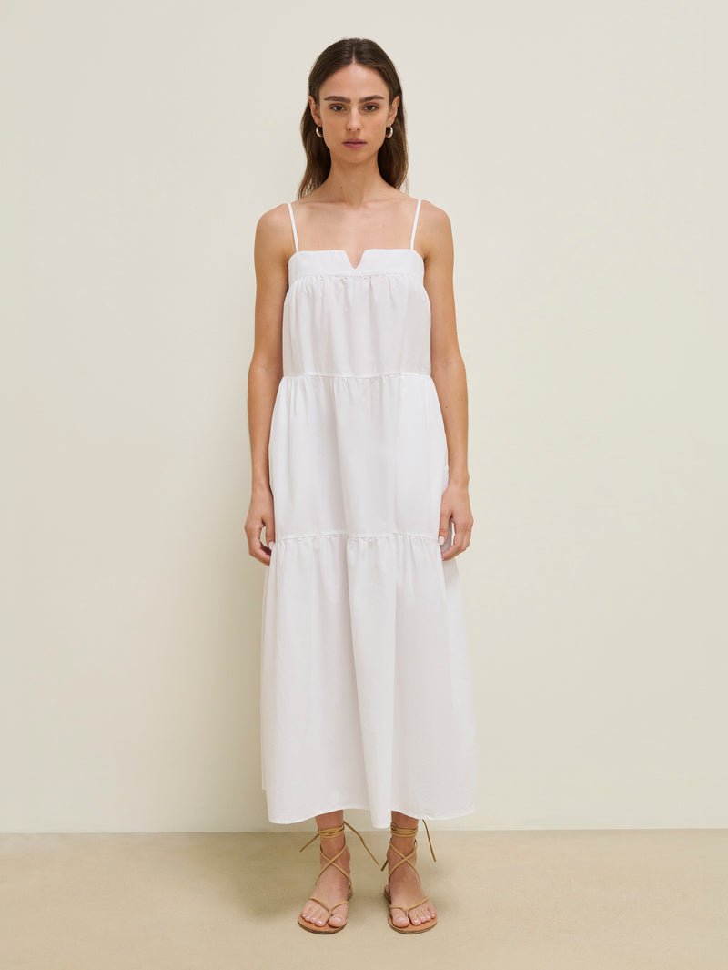 Woman in a white cotton midi dress and tan sandals stands against a plain background.