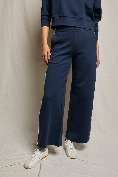 Model in navy wide-leg cotton pants and white sneakers, standing before a neutral fabric backdrop.