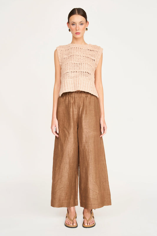 Woman in sleeveless beige knit top, brown relaxed-fit pants, and sandals stands against a plain backdrop.