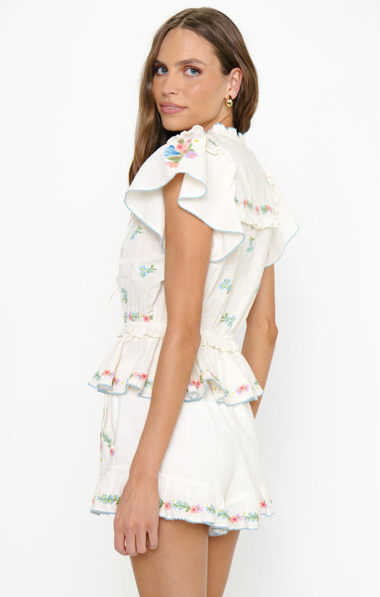 Woman in white embroidered shorts, lightweight fabric, casual wear, standing sideways and glancing over her shoulder.