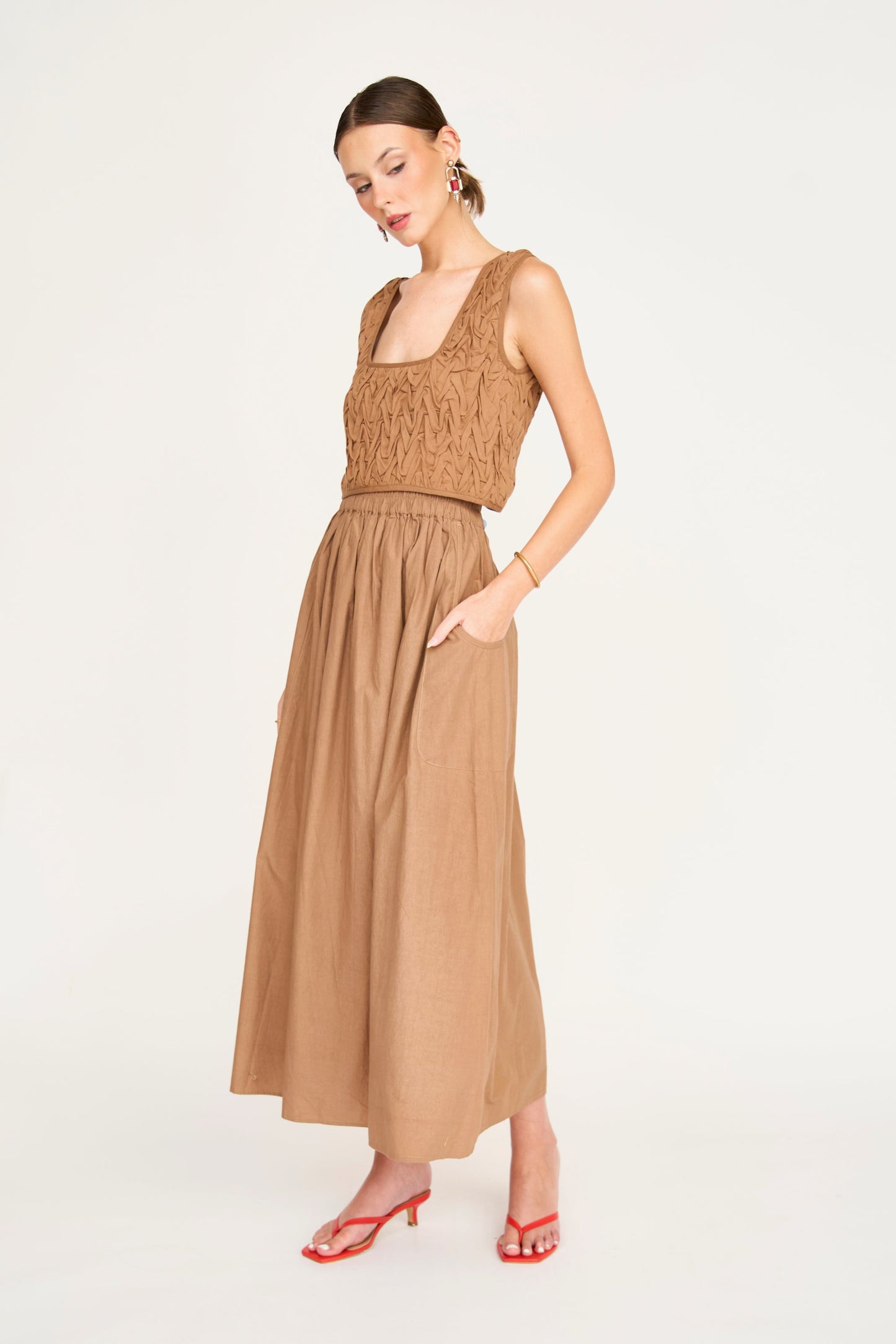 Woman in brown smocked top and matching skirt with red heels, standing against a white background.