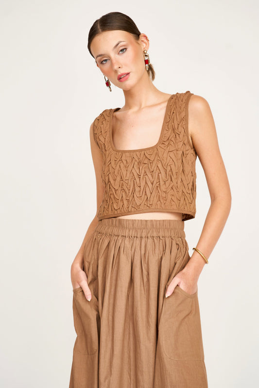 Woman wearing a brown smocked top with hands in pockets, standing against a plain background.