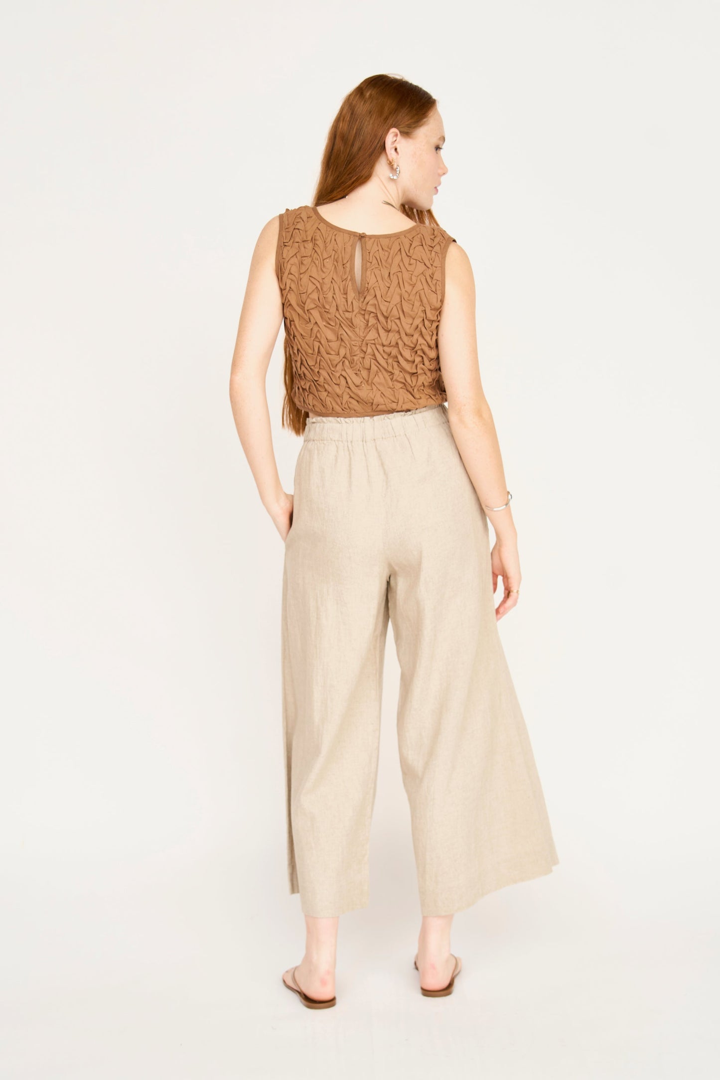 Woman shown from the back, in a brown smocked top, beige wide-leg pants, and sandals against a plain background.