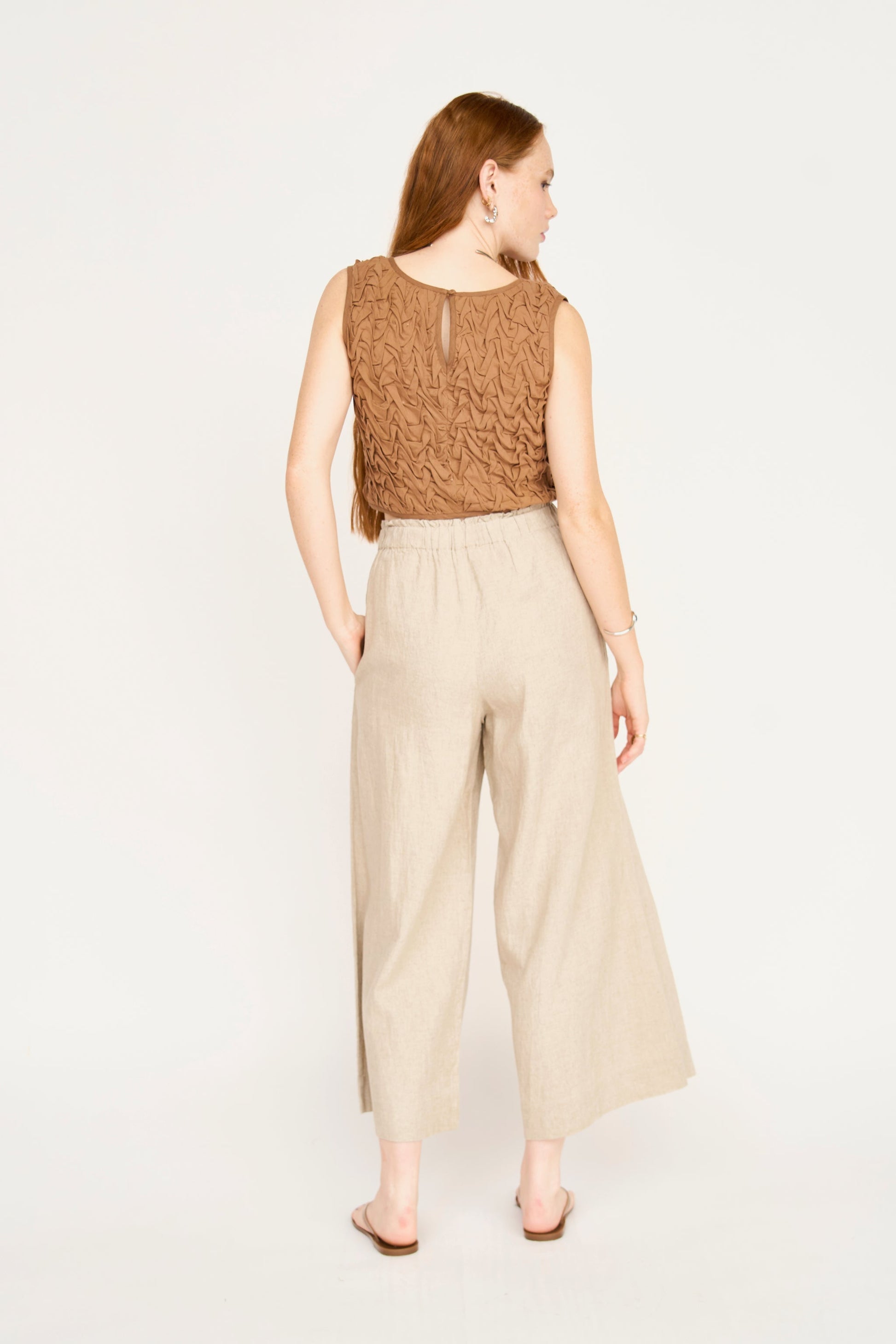 Woman shown from the back, in a brown smocked top, beige wide-leg pants, and sandals against a plain background.