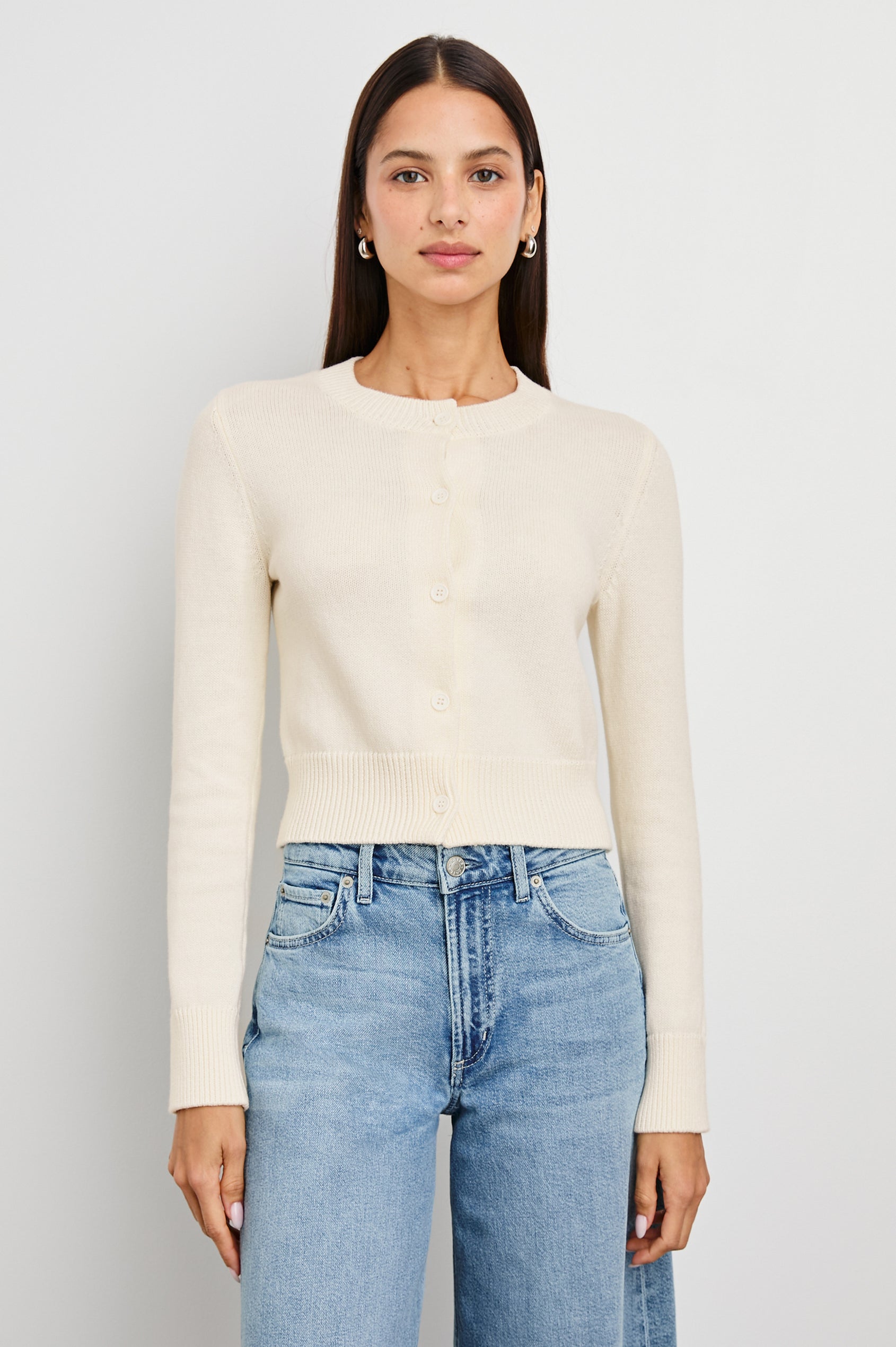 Woman in ivory knit cardigan and blue jeans stands against a white background, showcasing a versatile layering piece.