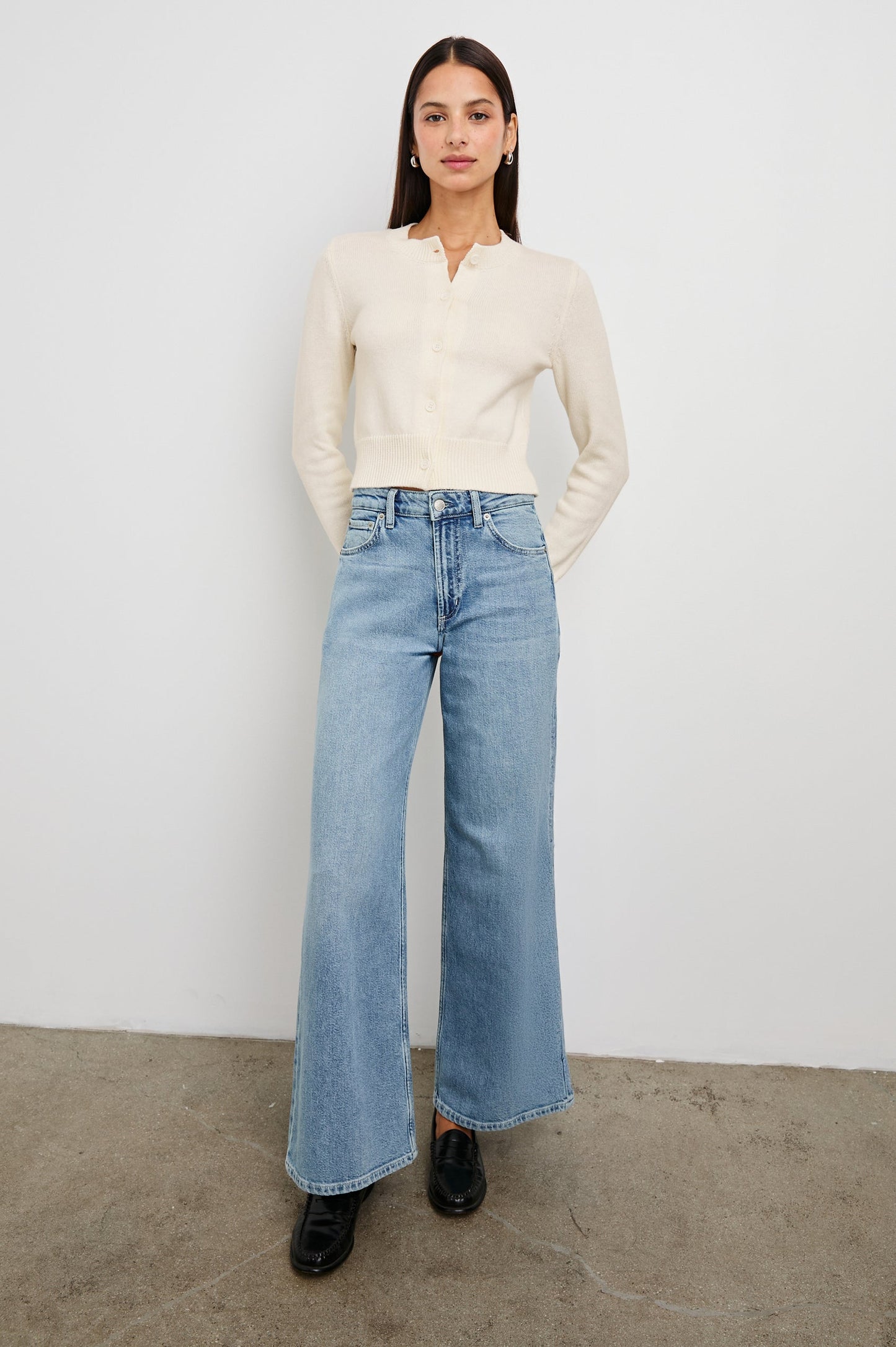 Ivory knit cardigan paired with wide-leg blue jeans; modeled standing against a white wall on concrete.