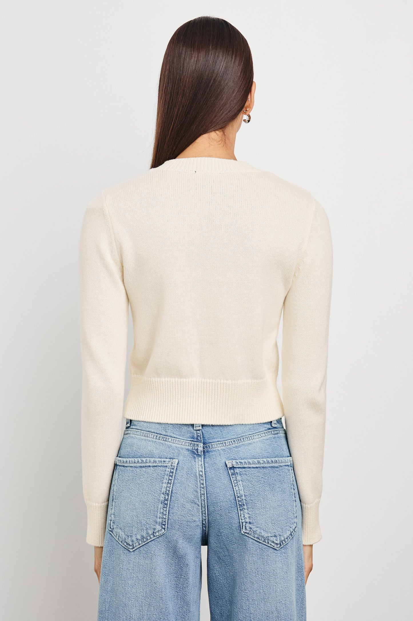 Back view of a woman wearing an ivory knit cardigan and blue jeans, standing against a plain white background.