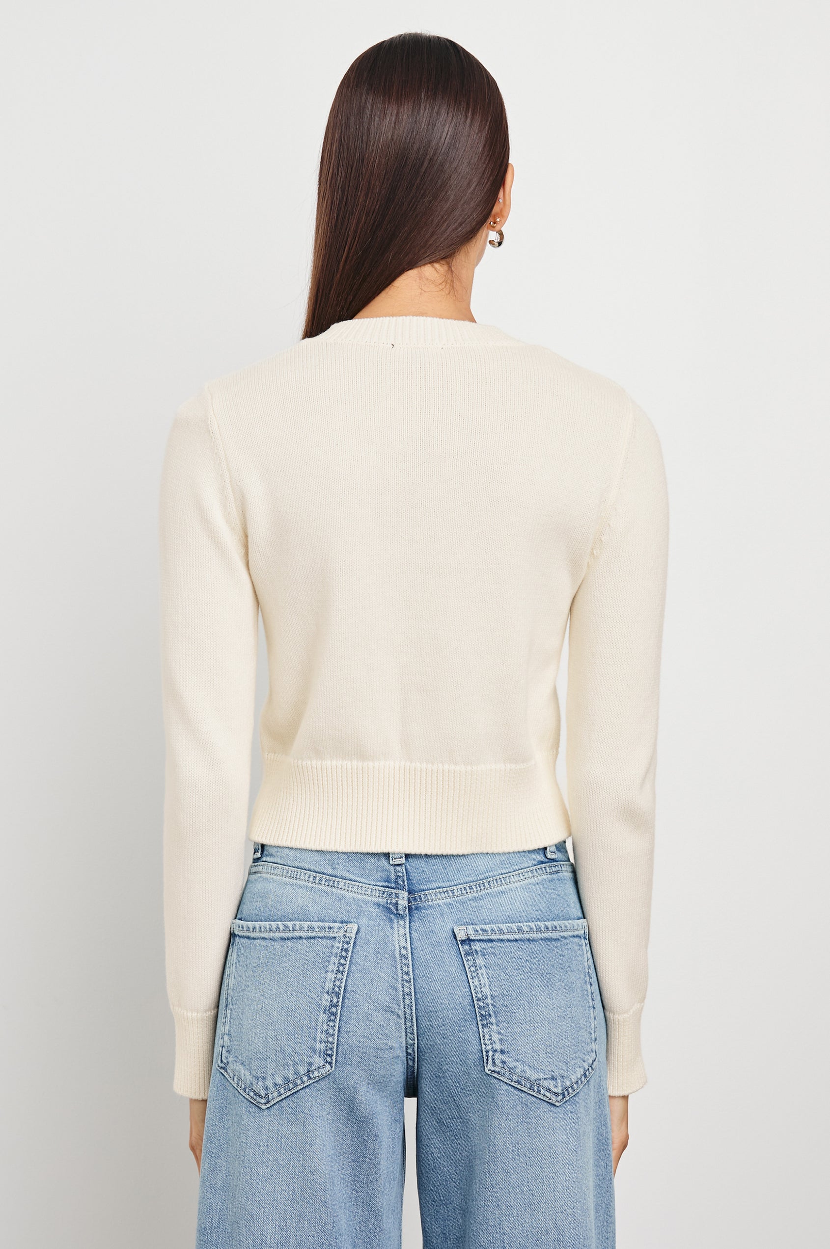Back view of a woman wearing an ivory knit cardigan and blue jeans, standing against a plain white background.
