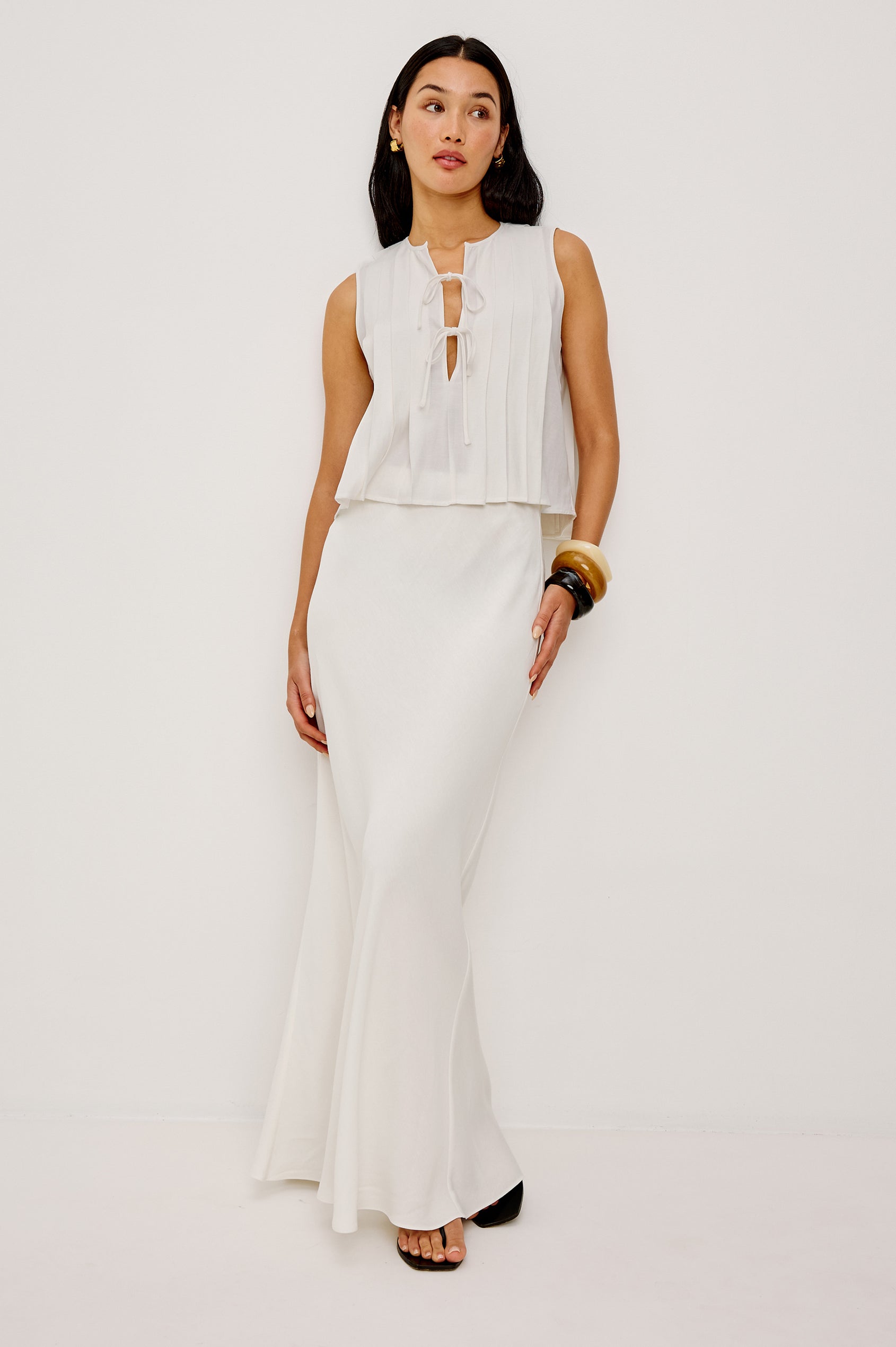Woman in ivory midi skirt and sleeveless white tie-front top stands against a plain white background.