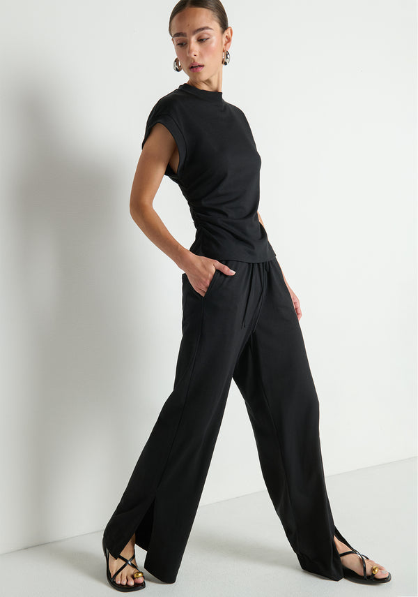Woman in black Rosetta pants and a matching black top, standing against a plain white background.