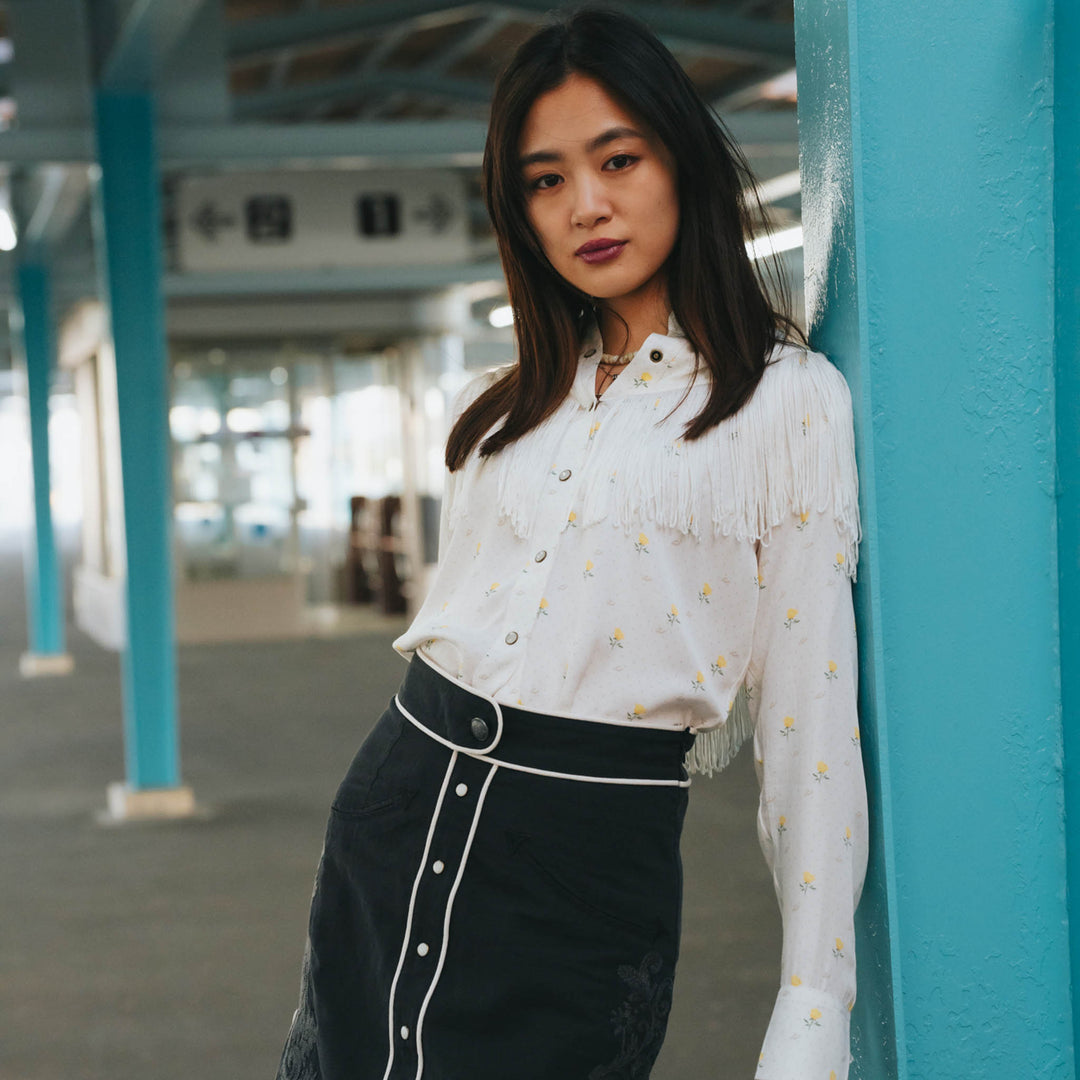 Woman indoors leans on blue pillar, wearing a short-sleeve blouse with red rose print, lightweight fabric.