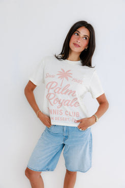 Woman in a white graphic tennis tee and denim shorts stands against a plain white background.