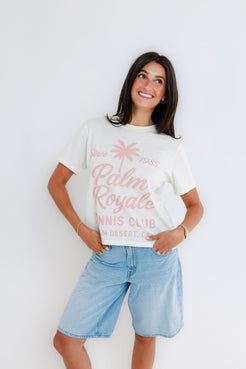 Woman in white cotton tennis tee and denim shorts smiles against a plain white background, showcasing casual wear.