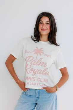 Woman in a white cotton tennis tee and blue jeans stands smiling against a plain background.