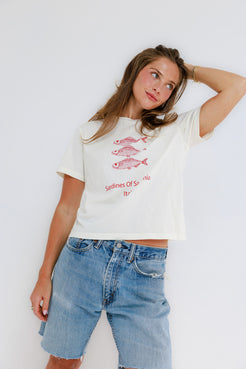 Woman in a white graphic cotton tee and blue denim shorts standing by a plain white wall.