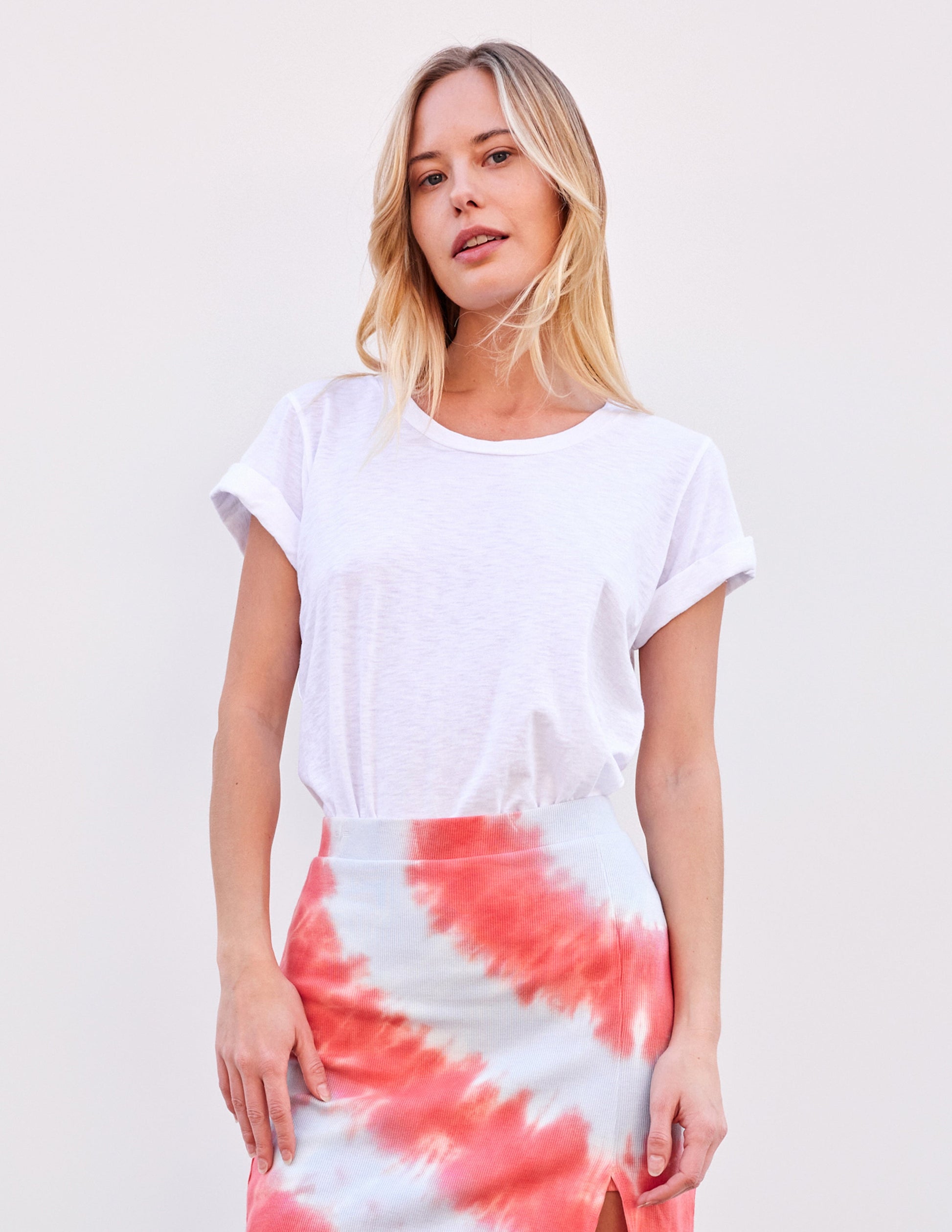 Woman wearing optic white rolled sleeve tee and pink tie-dye skirt, standing in front of a plain light background.