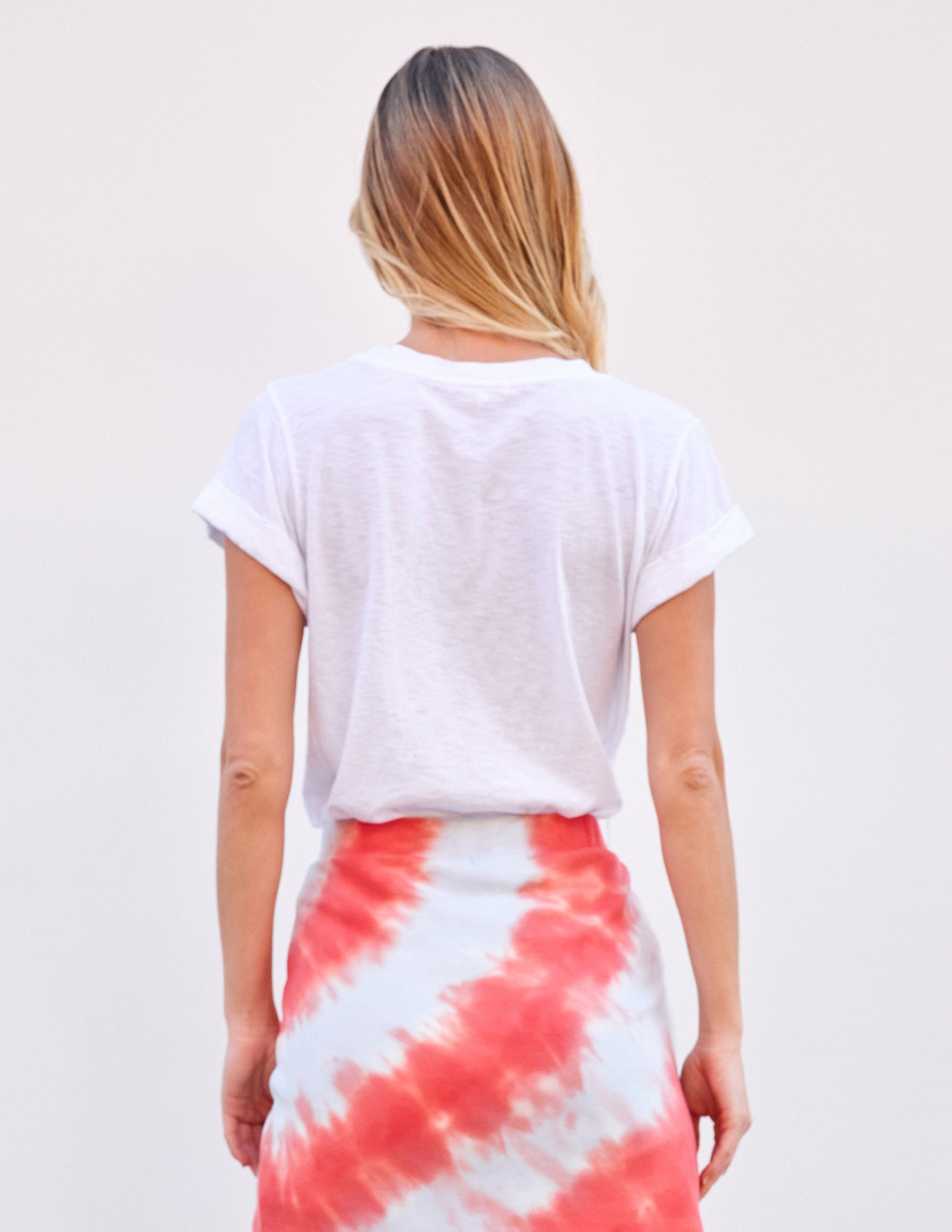 Back view of a woman in a white rolled sleeve tee and red-white tie-dye skirt against a plain light background.
