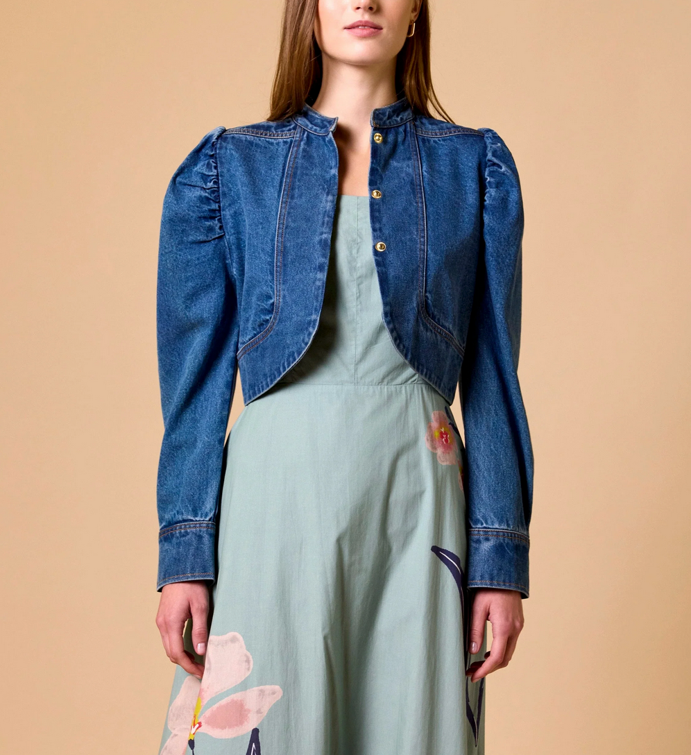 Woman in medium denim jacket over a light green floral dress, standing against a beige background.