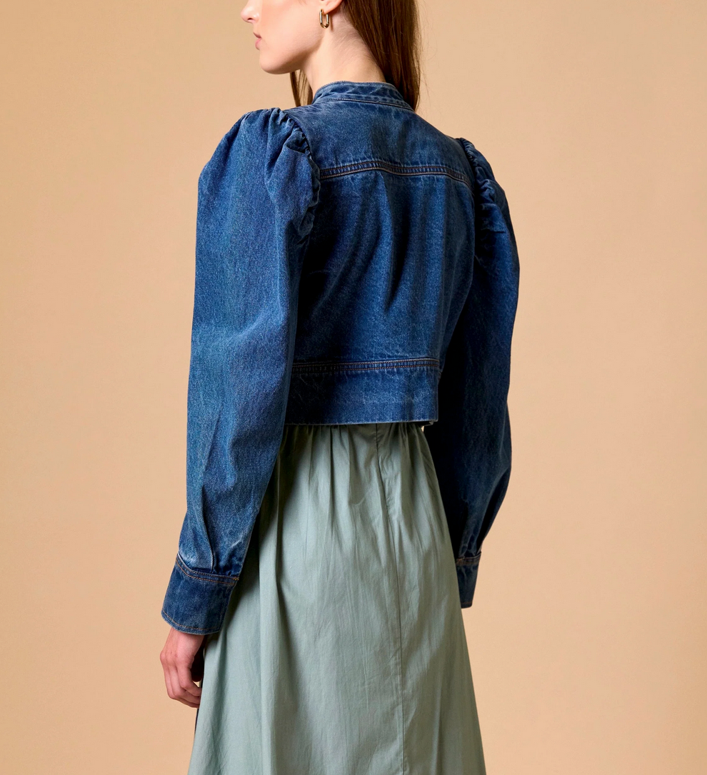Woman in medium denim jacket over sage green dress, standing against a beige background.