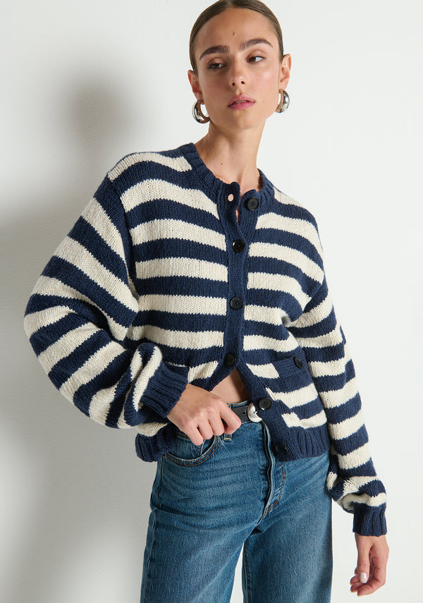 Woman in striped knit cardigan and blue jeans stands against a plain white wall.