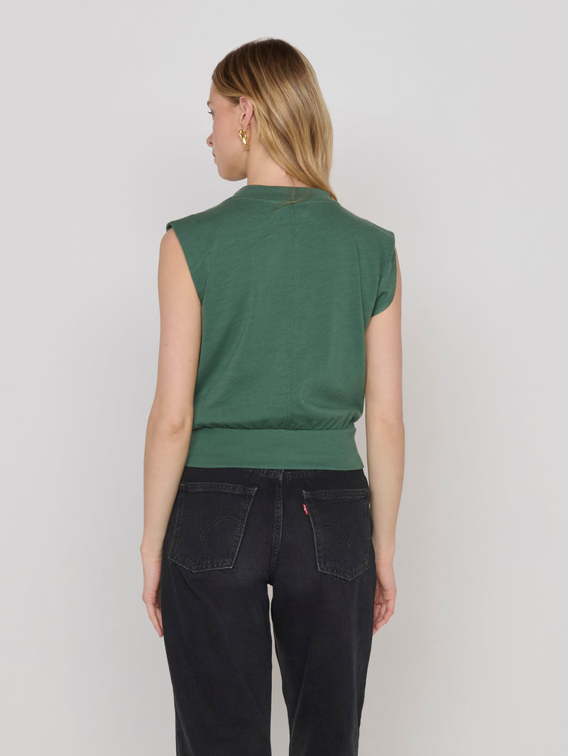 Woman in floral blouse and black jeans, facing away, against plain light background.