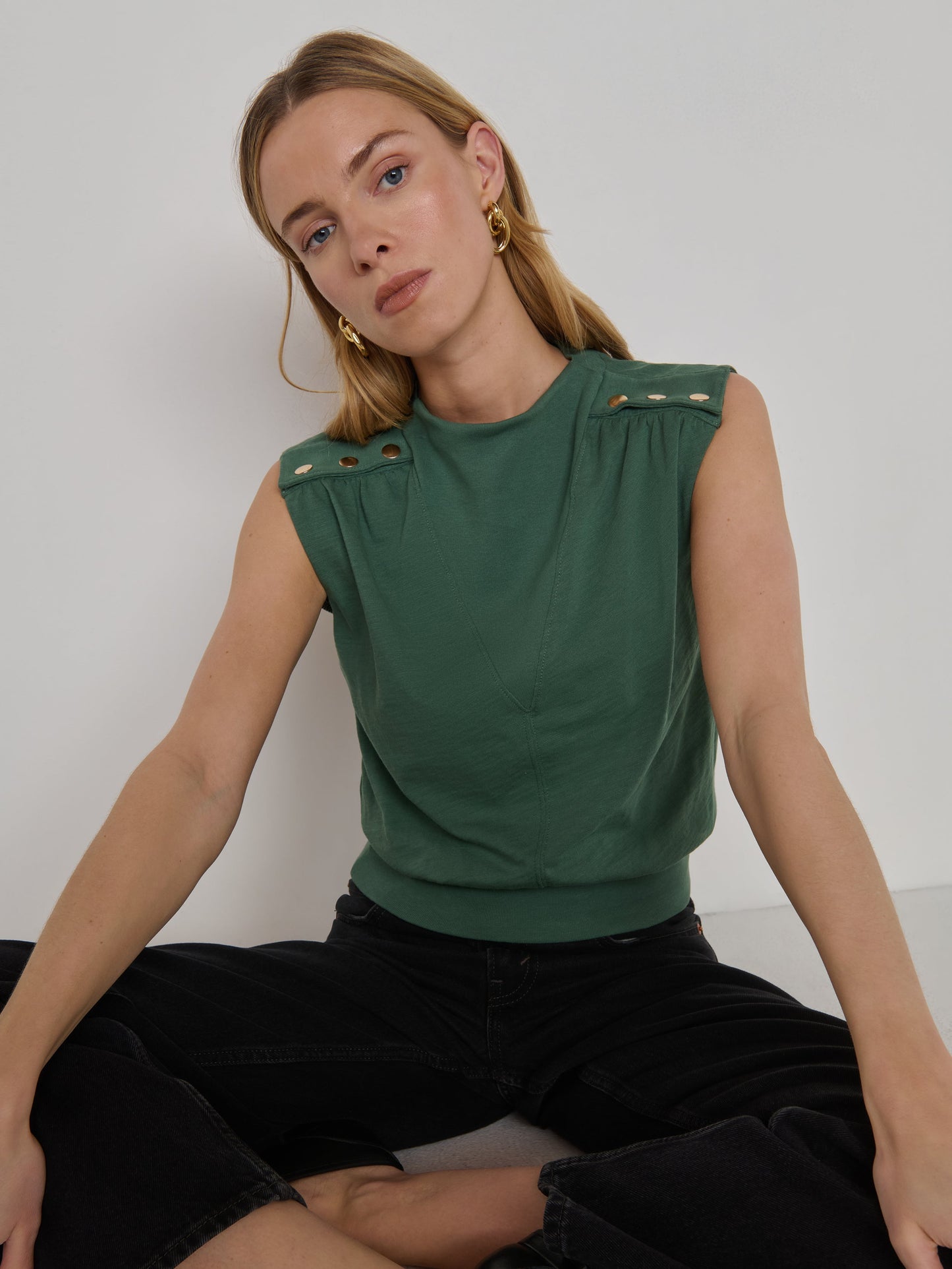 Woman in a sleeveless floral top and black pants sits cross-legged against a plain white background.
