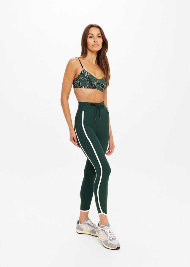 Woman in studio wears green recycled sports bra and leggings with gray sneakers, suitable for active or casual wear.