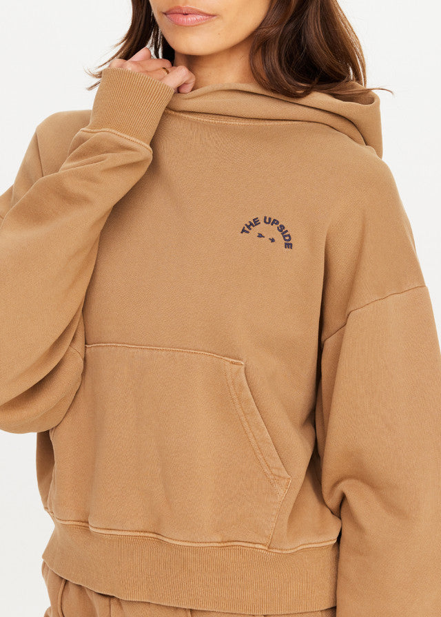 Woman in chai-colored hoodie with chest logo, soft material; one hand raised near chin.