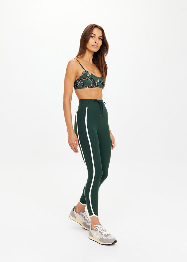 Woman wears green midi athletic pants, matching sports bra, and sneakers; outfit is suitable for workouts. White background.