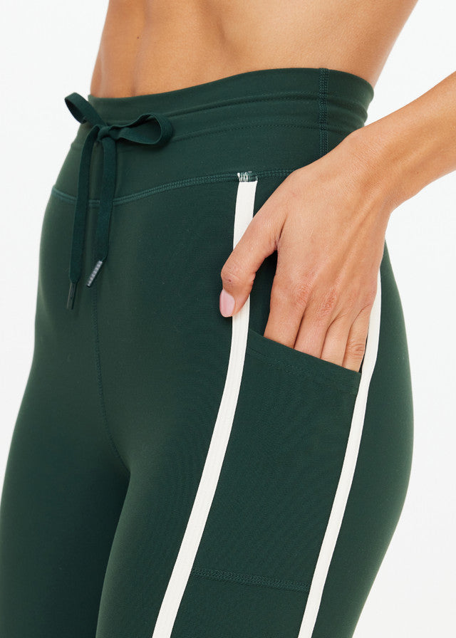 Woman in green midi pants with white side stripes, cotton-blend material, hand in pocket, shown from waist down.
