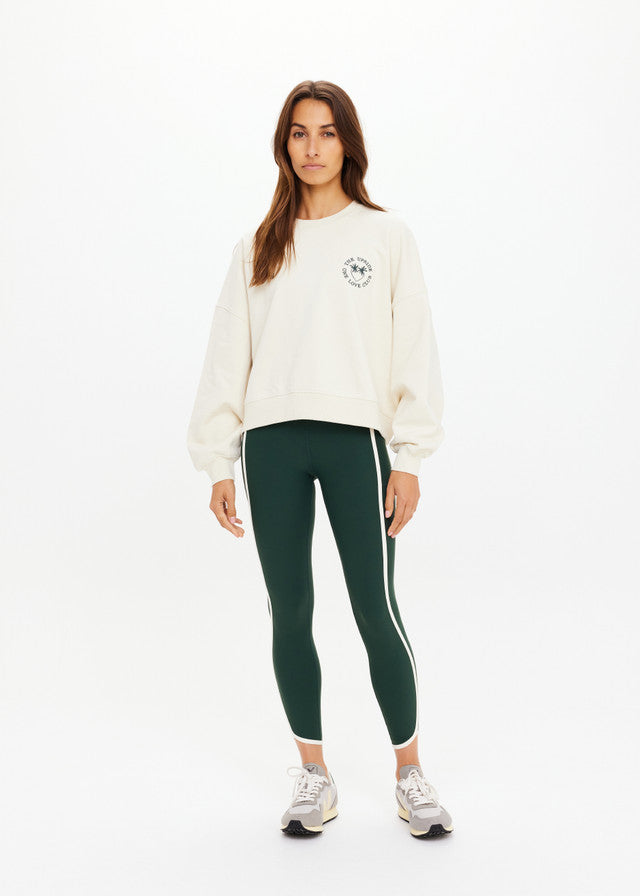 Woman in cream crewneck sweatshirt, green leggings with white stripes, and sneakers stands against a plain background.