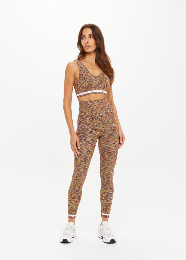 Woman wearing seamless black midi leggings, multicolored sports bra, and white sneakers, shown against a plain backdrop.