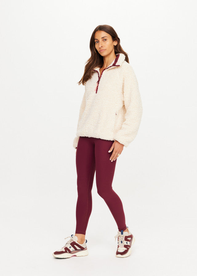 Woman in macadamia fleece pullover, burgundy leggings, and sneakers, standing against a plain white background.