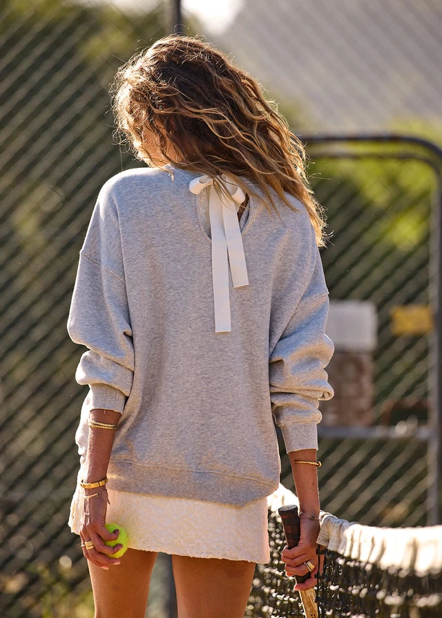Woman with wavy hair wears a grey marble crew sweater and white skirt on a tennis court.