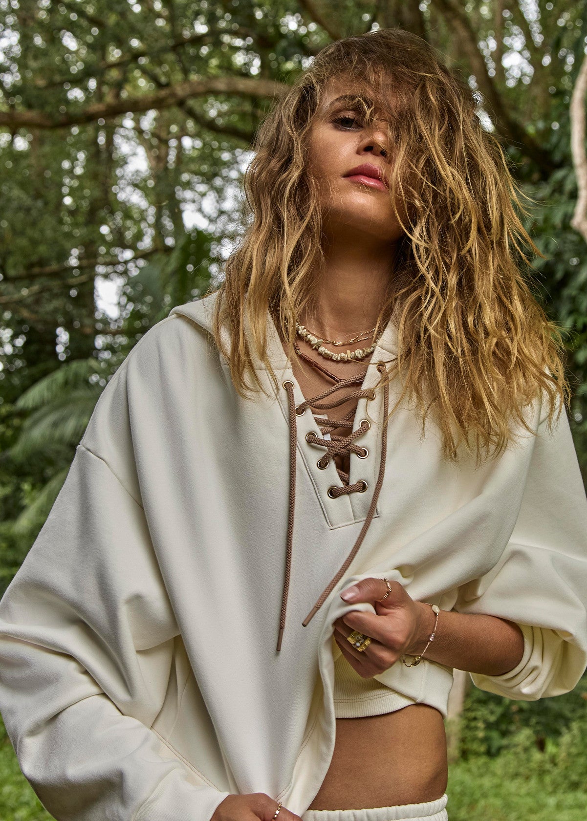 Woman with wavy hair in a light hooded cotton sweatshirt, standing outdoors among green trees and plants.
