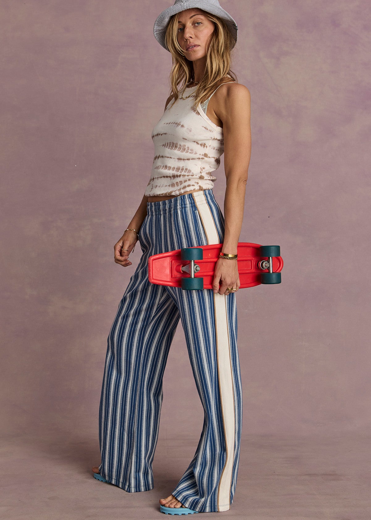 Woman in denim wide-leg pants, tie-dye tank, and hat holding a red skateboard against a purple background.