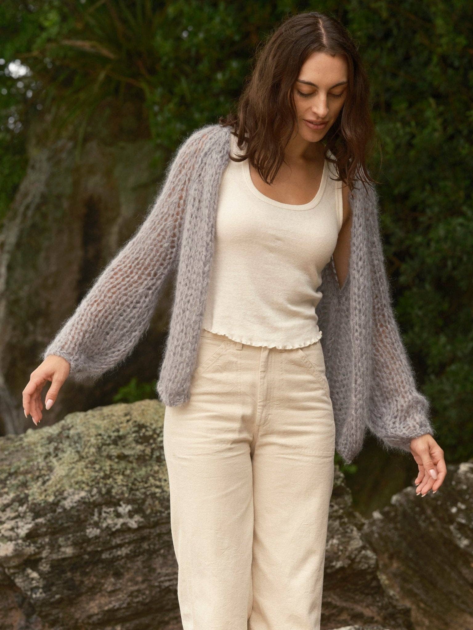 Woman outside wearing a knit cloud cardigan, cream tank top, and beige pants, looking down and smiling.