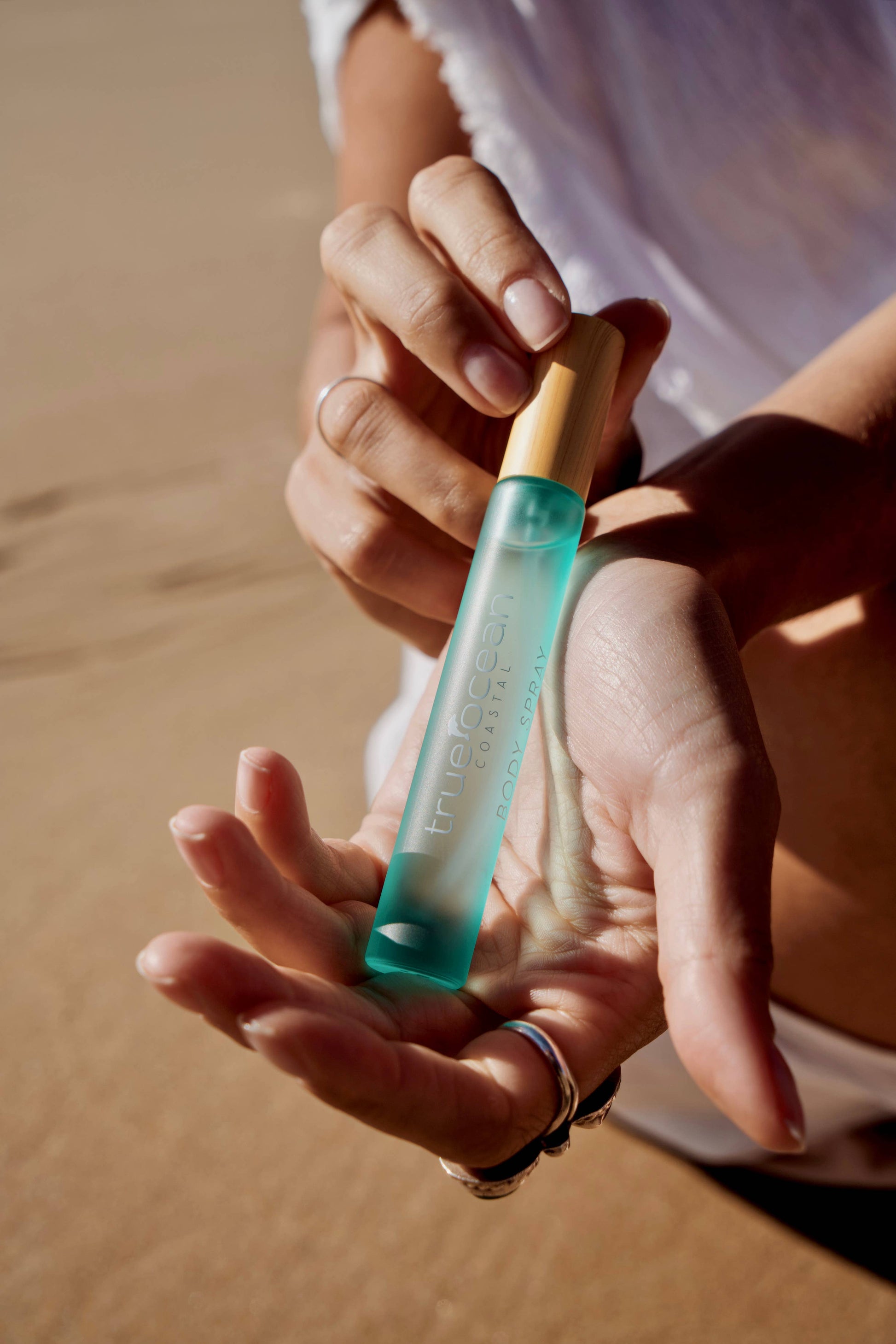 Person applies clear blue body spray to wrist at the beach, sunlight reflecting off their hand.
