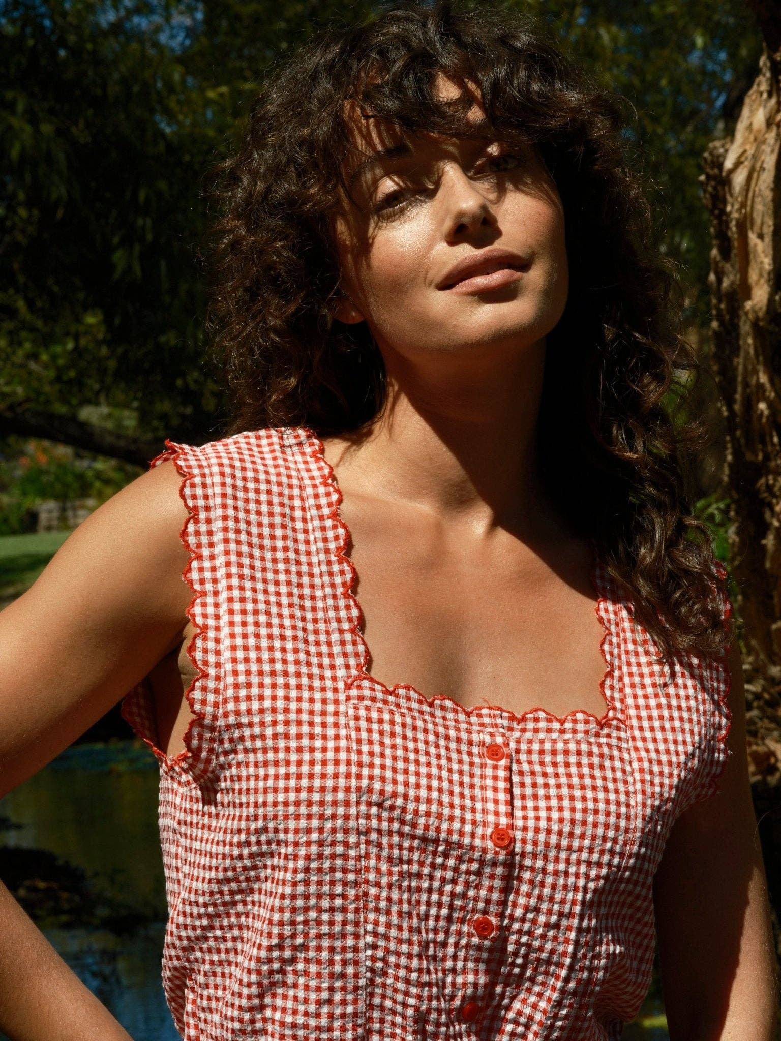 Woman with curly hair wears a sleeveless gingham cotton top with square neck, standing by a sunlit tree.